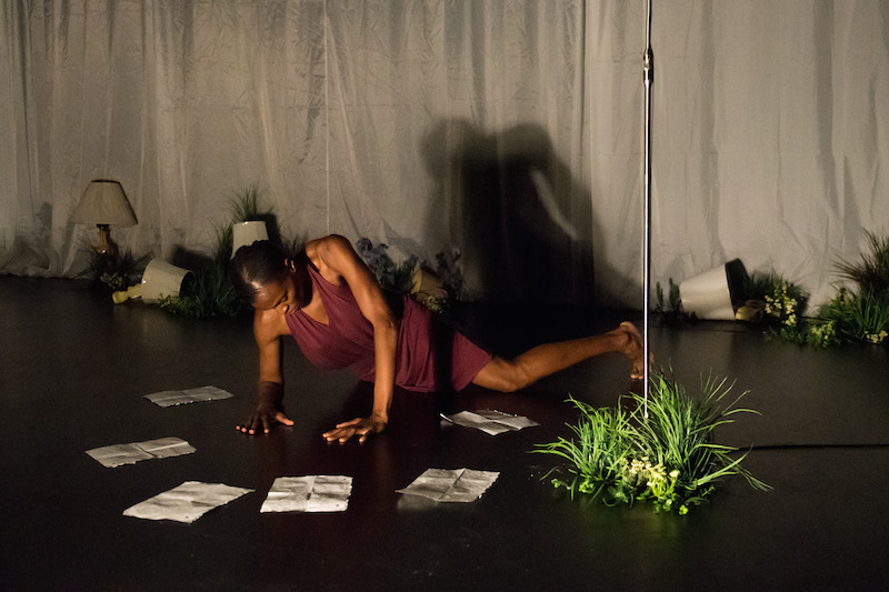 Okwui lays on the ground in a magenta halter dress. Notebook paper and plants litter the flower. Lamps are haphazardly tipped over.